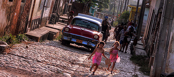 Girls running down the street and an old car in Cuba